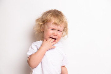 beautiful curly blond baby girl in in white t-shirt crying on a white background. the child has...