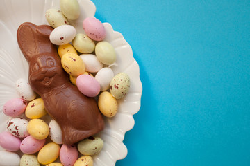 Easter chocolate bunny with small colorful eggs on a plate on a blue background. top view copy space