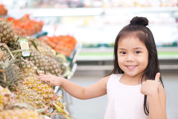 Asian child cute or kid girl smiling and take pineapple diet fruit with like and show thumb finger for buy or sale and food shopping in supermarket at department store on holiday