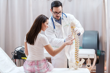 Female patient sitting with backs turned showing on spine model where she feel pain. Doctor holding...