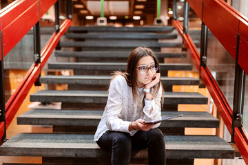 Bored collage girl with brown hair and eyeglasses leaning on the hand and sitting on the stairs and holding tablet. University building interior.