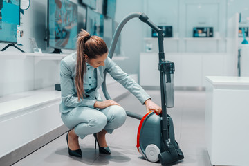 Happy Caucasian brunette in formal wear crouching and trying out new vacuum cleaner for her house.