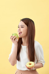 Choose a right choice for good health. Women is choosing choice between donut and green apple isolated over yellow.