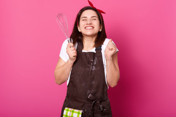 Smiling cheerful girl clenches fists with kitchen equipment in right hand, wears work clothing,...