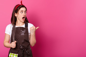 Surprised dark haired woman makes gesture with her thumb. Gifted active baker holds kitchen...