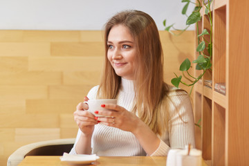 Obraz na płótnie Canvas Sincere cute young lady sits in local cafeteria near wooden shelf with green plant behind her, holds small white cup of coffee in both hands, looks in other way, warms up in cosy public place.