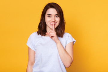 Body language concept. Beautiful smiling young female with happy expression, wears casual t shirt, demonstrates silence gesture, keeps fore finger on lips, isolated on yellow studio background.