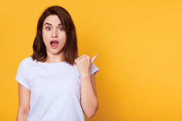 Close up portrait of beautiful brunette girl shows something aside with thumb, astonished woman points left and looks at camera with open mouth. Female poses confused over yellowbackground. copy space