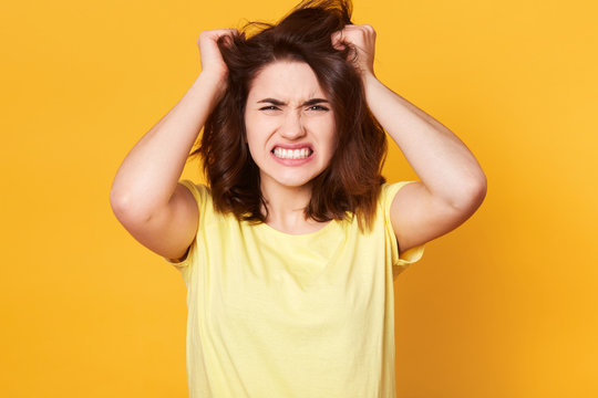 Attractive brunette female scrathes head and exclaims loudly, looks angry, wears casual t shirt, poses against yellow concrete wall. Emotive nervous woman worries because of faild examination.