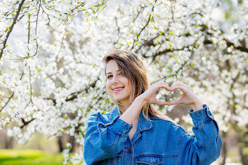 girl in a denim jacket stands near a flowering tree and show heart hand sign