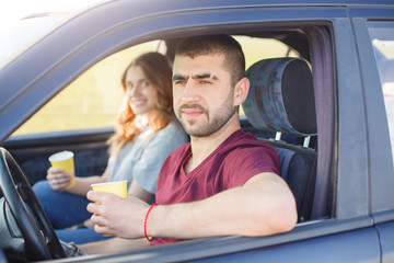Young male and female travel by car, family trip, couple spend time together during vacation, man and woman stop at side of road and drink coffe, having rest from long hours on their way. Traveling.