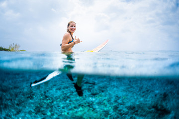 Happy woman sits on the surfboard in the ocean, looks at camera and smiles. Splitted image with underwater view