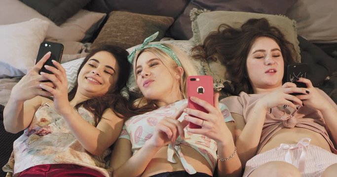 Pretty teenager ladies in a homewear clothes using a smartphone while laying down on the bed, eating chewing gum and feeling happy.
