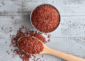 Bowl and full spoon with uncooked red rice on wooden background, top view