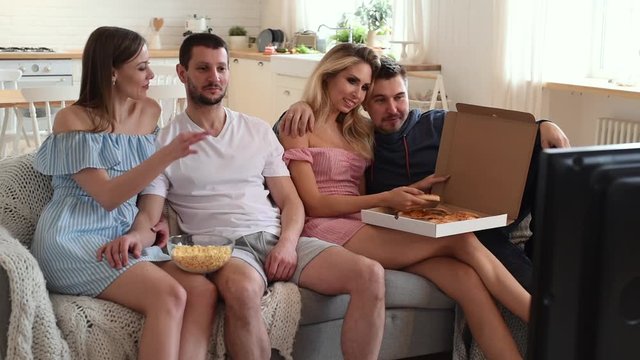 Group of friends watching TV together and eating pizza at home on sofa