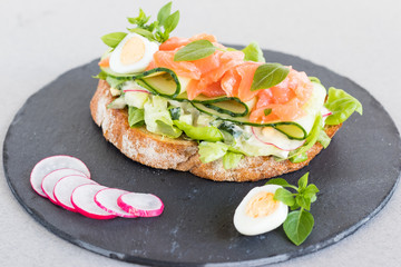 Danish open sandwich Smorrebrod with salmon on rye bread with vegetables and herbs on shale plate. Close up