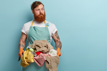 Busy male works about house, holds basin with clean towels and bottle of detergent, wears blue...