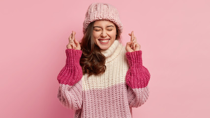 Hopeful optimistic young lovely woman with glad expression, makes wish to accomplish goals, raises hands with crossed fingers, wears oversized winter jumper and hat, isolated over pink background.