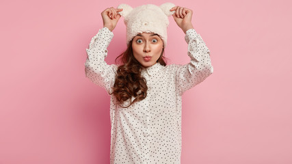 Horizontal shot of attractive European woman has folded lips, wears fashionable polka dot shirt and fluffy white headgear, keeps hands on ears, isolated over pink background. People and style concept