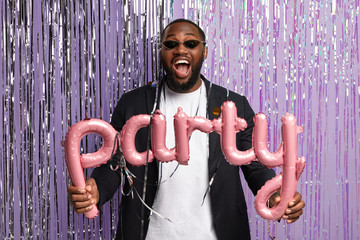 Amazing cheerful dark skinned guy in fashionable suit and sunglasses holds balloon letters meaning...