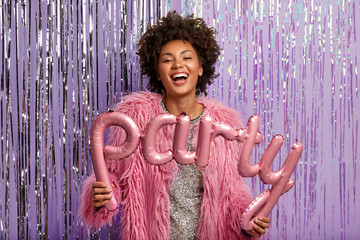 Glad dark skinned woman wears pink fluffy pink coat, holds allphabet balloons forming words party,...