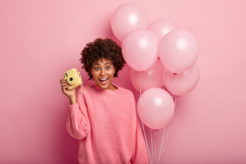 Photo of happy Afro American woman in oversized jumper, holds helium balloons, camera for making funny pictures, models over pink background. Festive event concept. Girl comes to congratulate friend