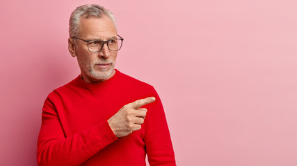 Photo of serious unshaven mature male demonstrates something aside on blank space, points fore...