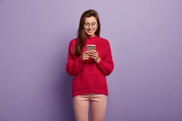 Photo of smiling Caucasian teenage girl focused in modern cell phone, enjoys online communication, wears long knitted red sweater, sends text messages to friend, stands over purple background