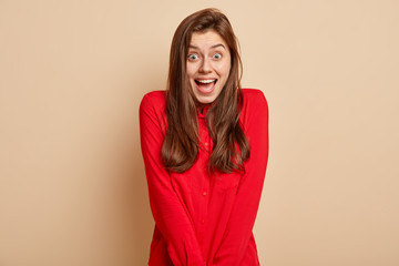 Headshot of dark haired young Caucasian woman with glad expression, expresses good emotions, wears red shirt, isolated over beige background, has toothy smile. People, happiness and joy concept