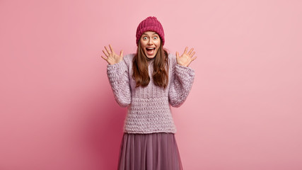 Nice happy young lady stares with eyes full of joy, raises hands and shows palms, wears knitted jumper and long skirt, feels excited, models over pink background. People, emotions, impressions