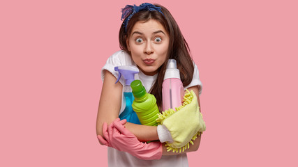 Headshot of happy surprised woman embraces bottles with detergent spray, holds mop, ready for cleaning her house, wears headband and white t shirt, models over rosy background. Hygiene concept