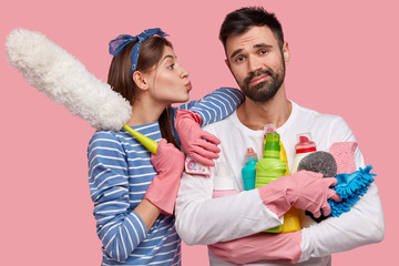 Indifferent fatigue unshaven man holds many bottles of detergent, his wife leans at shoulder, wants...