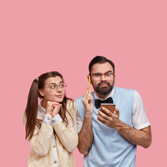 Photo of thoughtful man with thick bristle, holds notepad and pencil, thinks on ideas for writing essay, his girlfriend geek tries to help, stand closely to each other, work together, isolated on pink