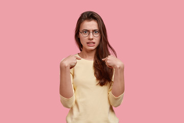 Photo of embarrassed lovely woman points at herself, has annoyed facial expression, wears transparent glasses and jumper, reacts emotionally on being blamed, stands indoor over pink background.