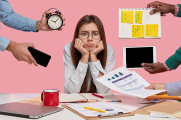 Sad female workaholic keeps hands under chin, busy making project work, studies papers, wears elegant white shirt, sits at desktop, unknown people stretch hands with notes, alarm clock, smartphone - obrazy, fototapety, plakaty