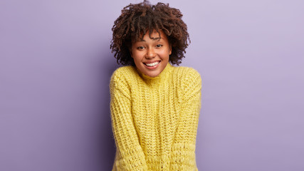 Horizontal shot of attractive dark skinned female has sincere smile, feels optimistic and glad, being in good mood, dressed in yellow jumper, poses against lilac studio wall. People and emotions