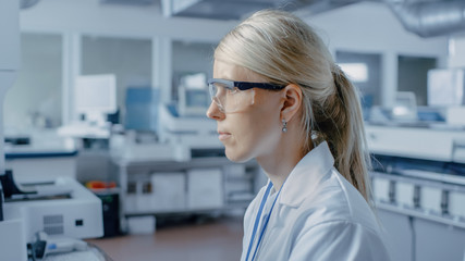 Portrait Shot of Female Scientist Sits at His Workplace in Laboratory, Uses Personal Computer. In the Background Genetics Research Centre with Innovative Equipment.