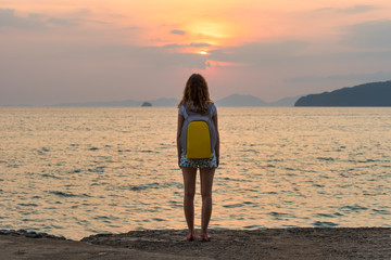 Fototapeta na wymiar One young female tourist traveler with a backpack stands and looks at the sea at the beautiful sunset and seascape. Full rear view.