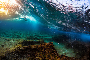 Underwater view of the ocean wave breaking over the shallow reef with sharp stones. Surfer floats on the background