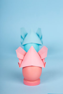 Happy Easter holiday concept dyed chicken pink and blue eggs in a bunny origami hats with ears on blue paper background with copy space. COVID-19 easter concept