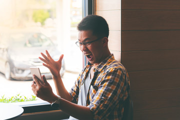 Asian guy Showing excitement and joy from the information received on the mobile phone, while sitting in a coffee shop, to technology communication concept.