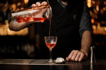 Male bartender pouring a delicious red cocktail from the measuring glass cup through the strainer