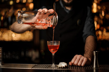 Bartender pouring a delicious red cocktail from the measuring glass cup through the strainer