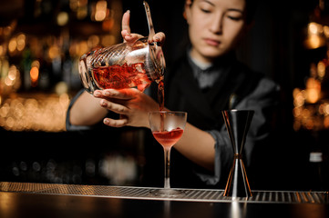 Bartender girl pouring a delicious red cocktail from the measuring glass cup through the strainer