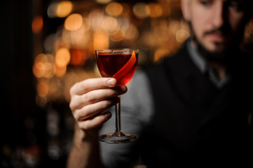 Bartender serving a transparent red cocktail in the glass decorated with a lemon zest on the steel bar counter