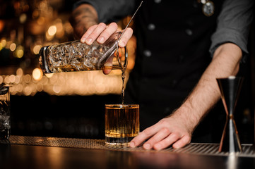 Bartender pouring an alcoholic drink from the measuring glass cup through the strainer to the cocktail