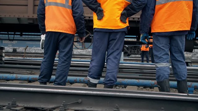 Railway workers are standing and looking at a passing train. Legs close-up. Worker in orange workwear. In the background traveling freight cars