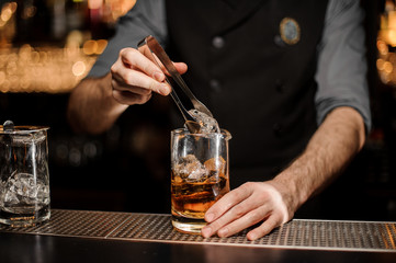 Professional bartender adding a big ice cube to the measuring glass cup with a brown cocktail