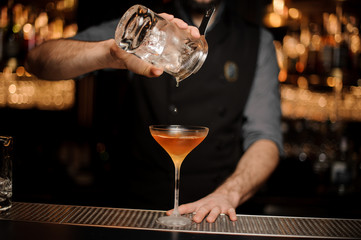 Bartender pouring an alcoholic drink from the measuring cup through the strainer to the cocktail glass