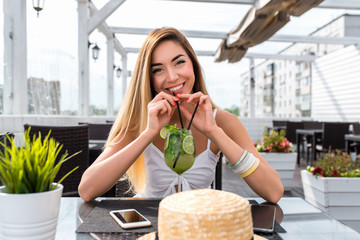 The girl in summer cafe on porch, happy smiling laughs drinking a cocktail through a straw. Relax in restaurant after work, long hair white dress. Business lady at dinner, emotions of joy and fun.